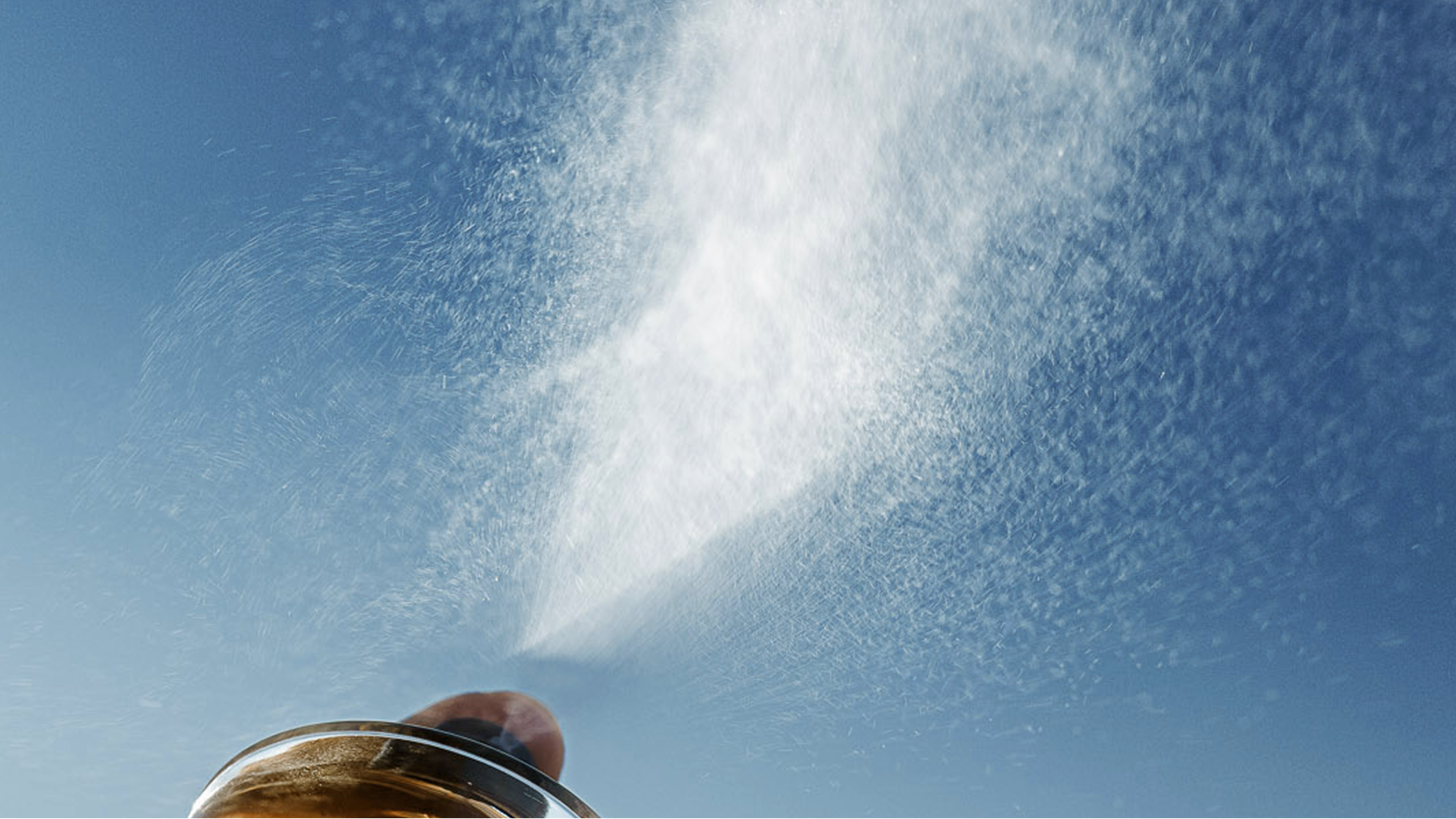 A close-up low-angle shot of a metallic sprinkler head spraying a fine white water mist against a clear blue sky.