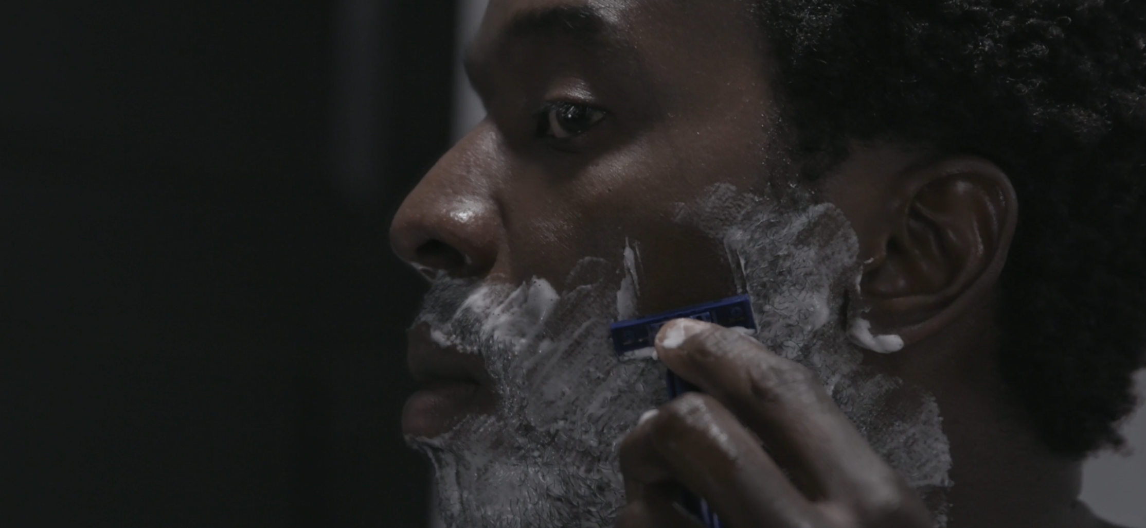 Man shaving with a blue razor and white shaving foam on his face, showcasing grooming routine.